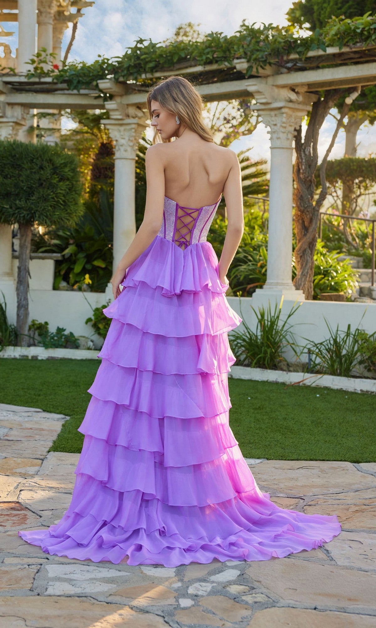 A woman stands outdoors on a stone path, facing away, wearing the Long Prom Dress: Juliet JT2738S, which features a strapless bodice and chiffon ruffle skirt with greenery and classical columns in the background.